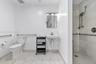 Bathroom featuring light-toned tile flooring, wall-mounted sink, walk-in shower with stone-finish wall tile, and metallic wall-mounted grab bars