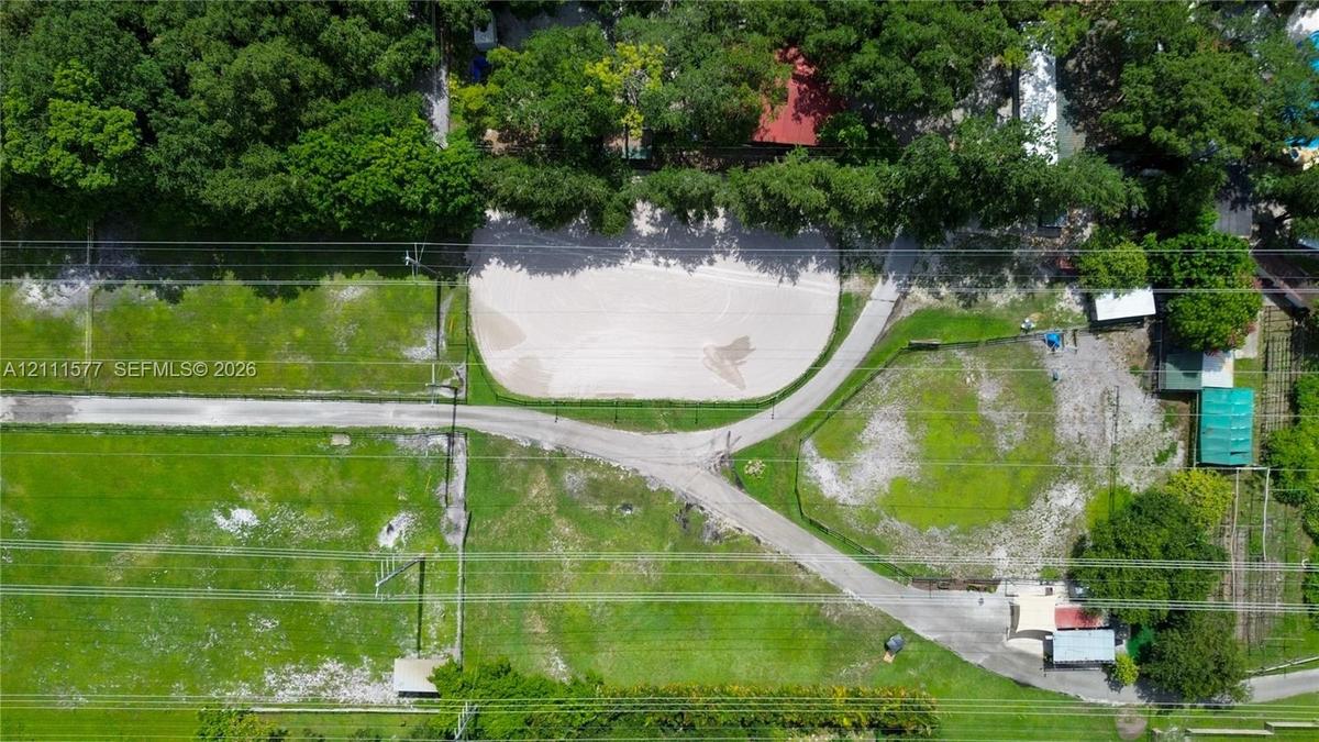 Aerial view of property featuring a sand-surfaced arena, cleared pasture land, winding paved pathways, and mature tree-lined perimeters