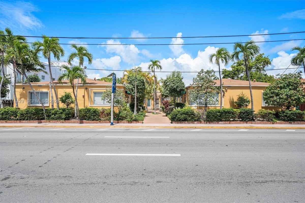 Street view of the low-rise building with a yellow stucco facade, palm trees and landscaped shrubbery