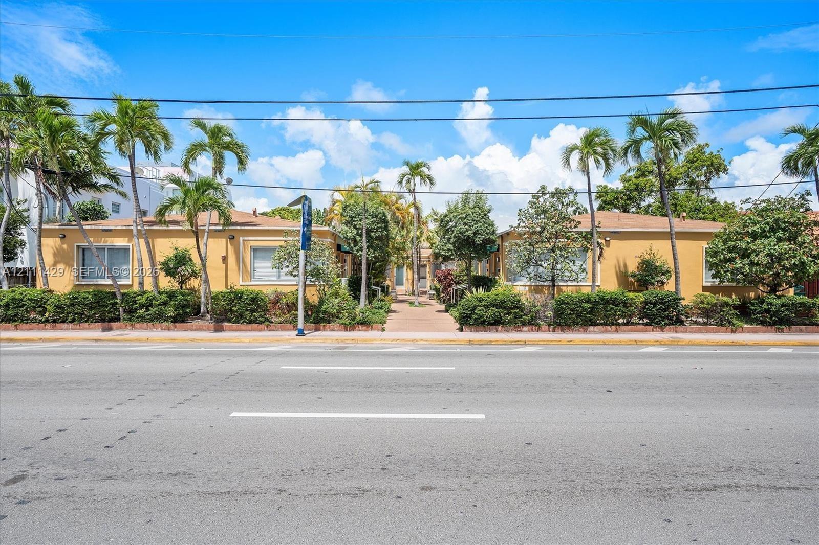 Street view of the low-rise building with a yellow stucco facade, palm trees and landscaped shrubbery