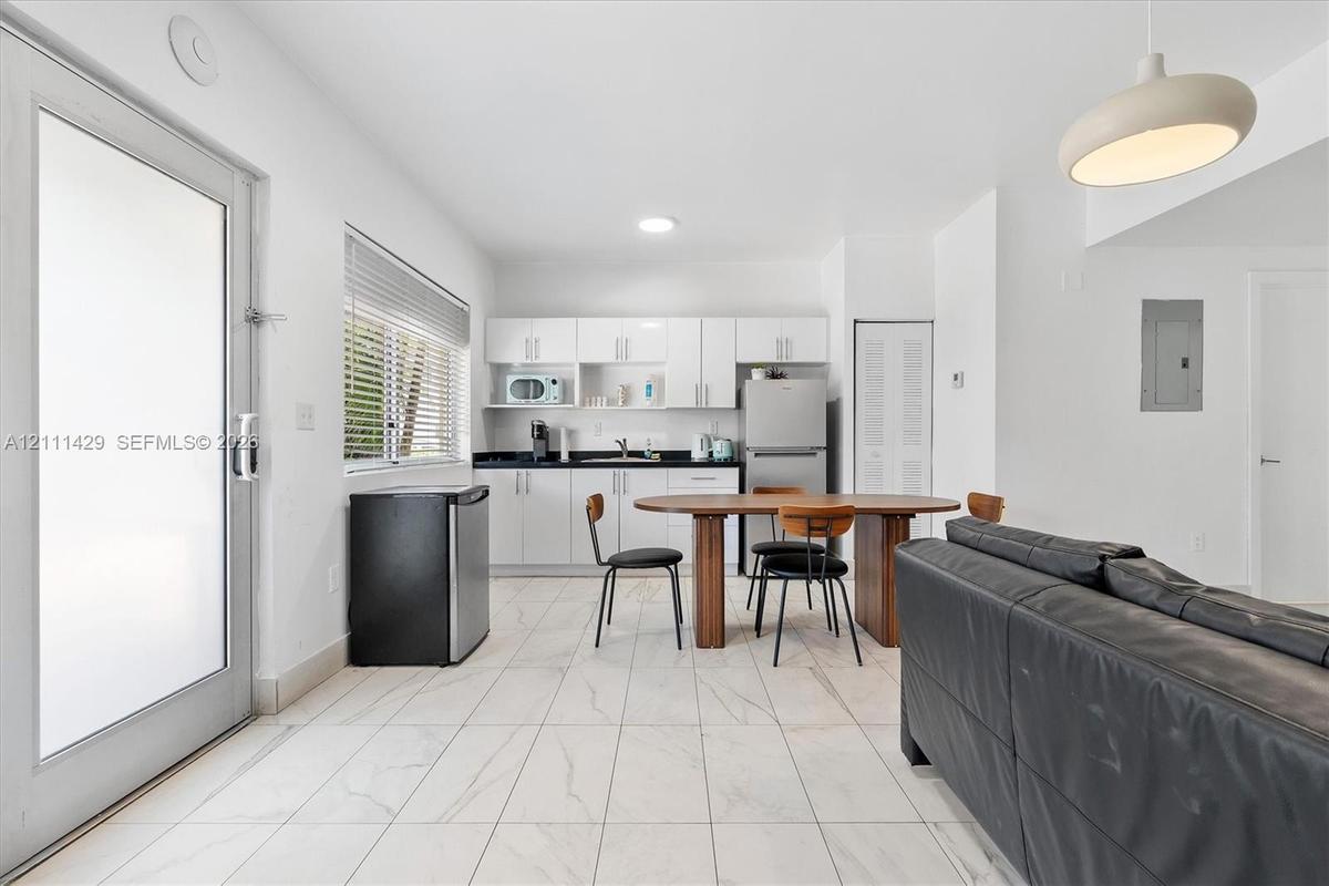 Open living and dining area with a wood dining table, black chairs, white kitchenette cabinetry, a stainless steel refrigerator and a louvered closet 