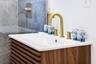 Slatted wood-finish vanity with an integrated white sink, topped by a widespread gold-tone faucet and matching hardware, set against blue-toned stone-