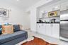 Living area with a sofa bed and large-format tile flooring, next to a kitchenette with white cabinetry, black countertops and a stainless steel refrig