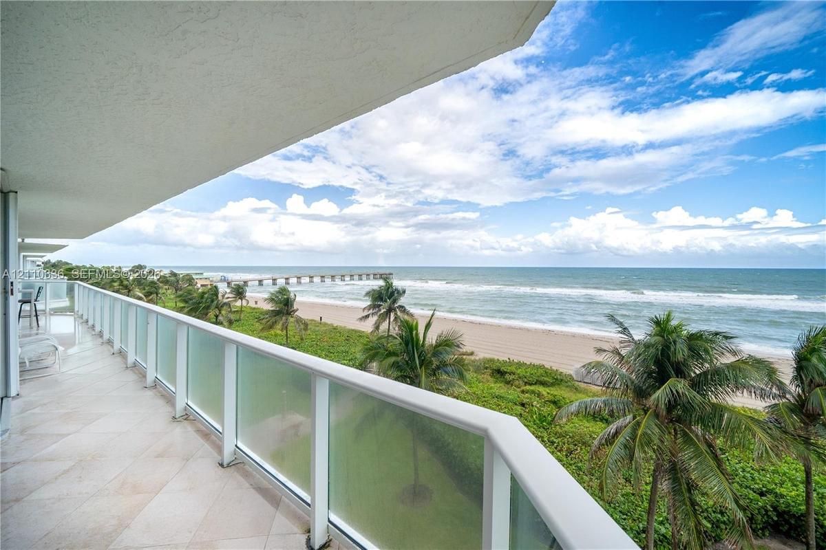Covered balcony featuring tile flooring and a glass-paneled railing overlooking the ocean, sandy beach, and palm trees