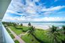 Elevated balcony overlooking coastal frontage featuring palm trees, manicured green space, paved walkways, and a pier extending into the ocean