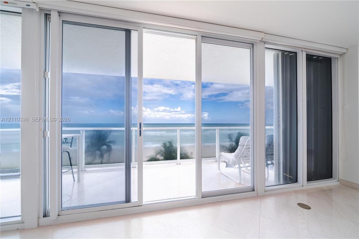 Floor-to-ceiling sliding glass panels, balcony with glass railing, stone flooring, and expansive ocean scenery