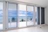 Floor-to-ceiling sliding glass panels, balcony with glass railing, stone flooring, and expansive ocean scenery