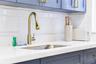Blue shaker-style cabinetry with brass-finish hardware, undermount stainless steel sink, gold-tone gooseneck pull-down faucet, white solid-surface cou