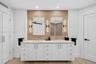 Dual-sink custom vanity featuring white cabinetry, light-toned stone counter, warm wood inspired wall tile and wall-mounted vertical sconces