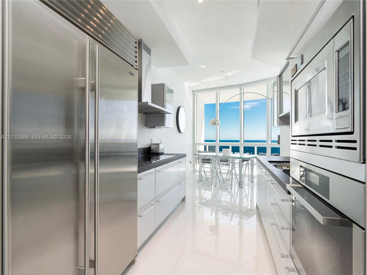 Modern galley kitchen featuring high-gloss cabinetry, stainless steel professional-grade appliances, dark stone countertops, polished tile flooring, a