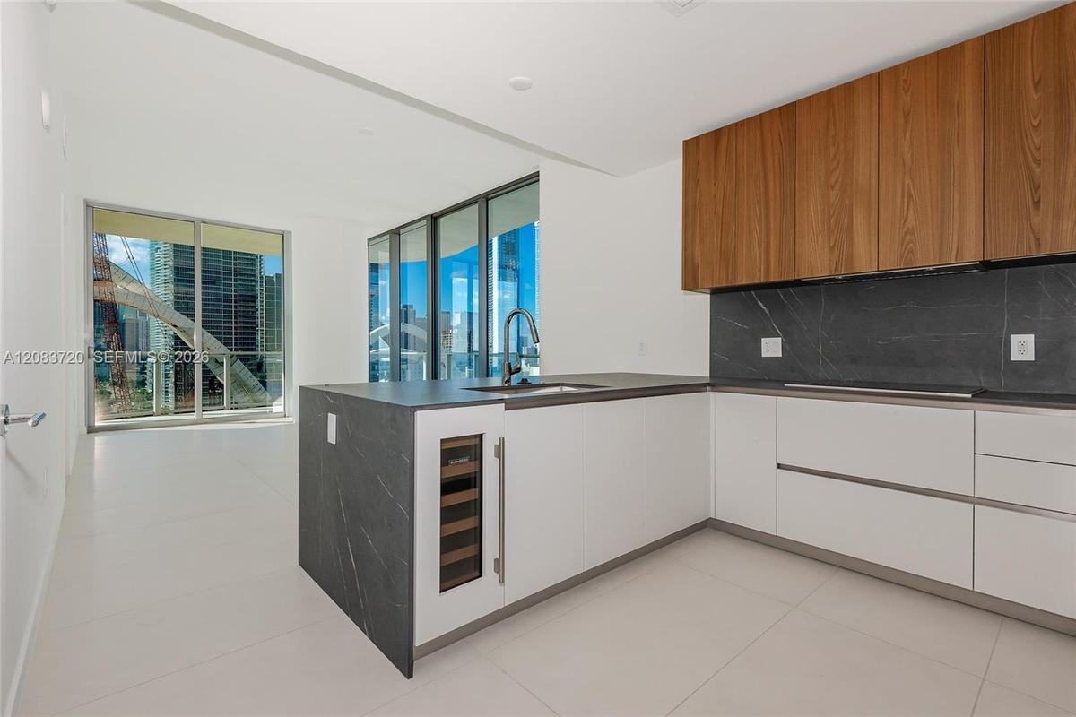 Modern kitchen featuring white flat-panel cabinetry, wood-veneer upper cabinets, integrated wine refrigerator, dark stone-finish countertops and backs
