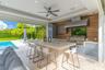 Covered outdoor kitchen featuring a stone-topped island, stainless steel grill, professional-grade range hood, wood-finish wall paneling, and recessed