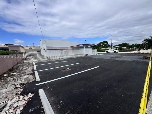Asphalt-paved parking area featuring marked spaces, concrete parking bumpers, a perimeter metal fence, and an exterior structure with a covered entran