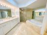 Dual-sink vanity with stone countertop, walk-in glass-enclosed shower, soaking tub with stone tile surround, stone flooring, and mirrored wall surface