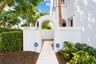 Stucco-finished exterior wall featuring an arched entryway, concrete walkway, wall-mounted exterior lighting, and landscaped perimeter hedges