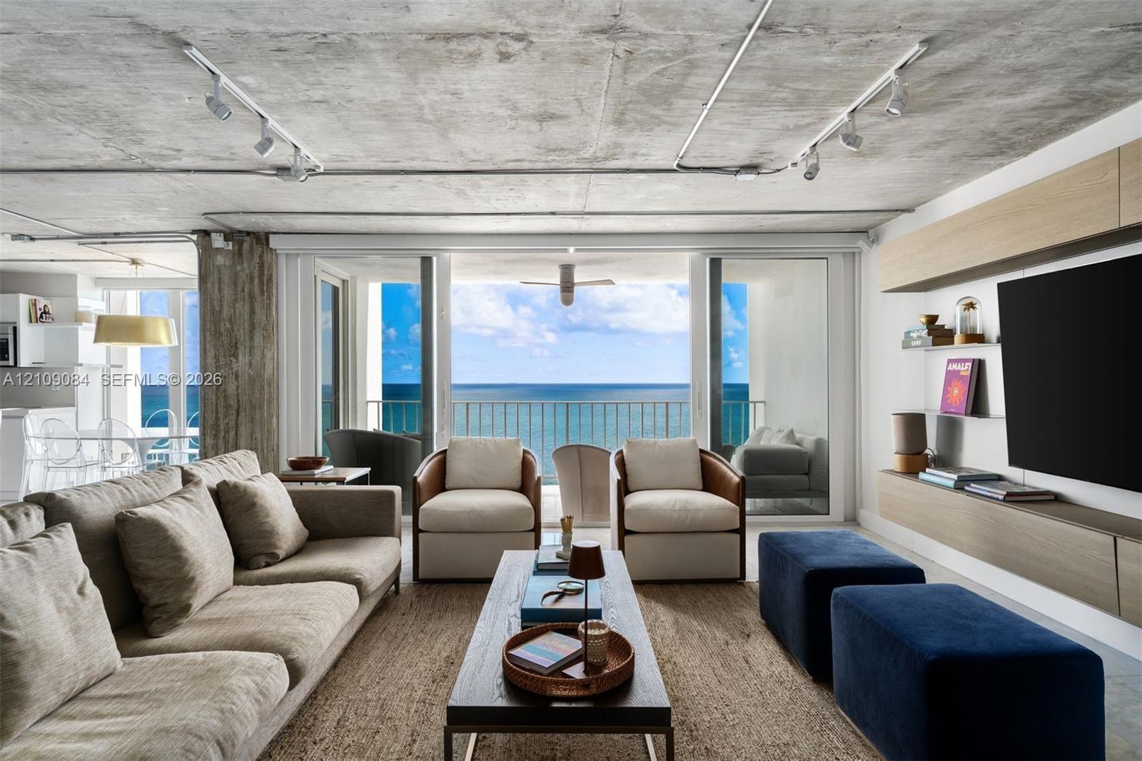 Exposed concrete ceiling with industrial track lighting, floor-to-ceiling glass doors, wall-mounted light wood-finish cabinetry, and balcony access fe