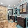 Bathroom featuring a walk-in shower with stone tile surround, dual dark-finish vanities with integrated cabinetry, beadboard wall paneling, and stone 