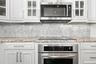 White shaker-style cabinetry with glass-front uppers, stone-finish countertops, mosaic tile backsplash, and stainless steel built-in appliances includ