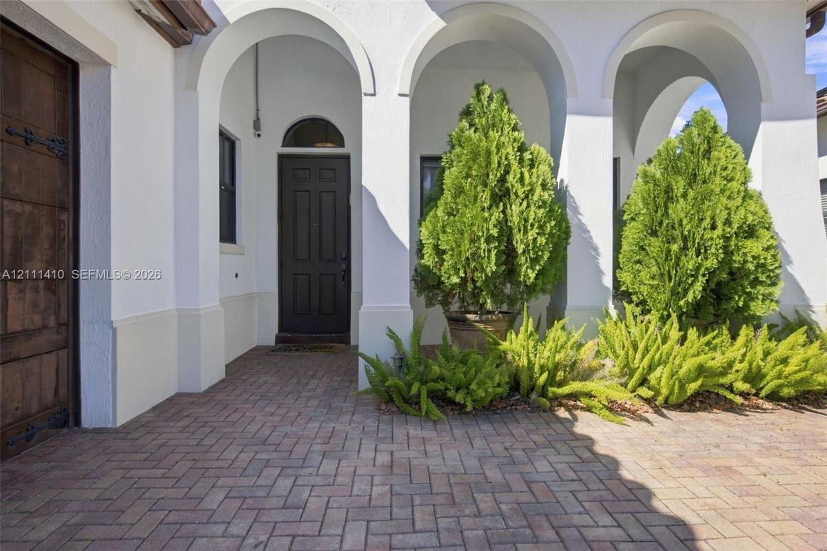 Covered entryway featuring arched architectural detailing, herringbone-patterned brick pavers, a solid wood-finish entry door with metal hardware, and