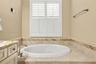 Built-in soaking tub with a tile surround, herringbone-patterned stone backsplash, plantation shutters, and a vanity featuring a stone countertop