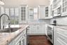Kitchen featuring custom white cabinetry with glass-front uppers, stone countertops, stainless steel appliances, a mosaic tile backsplash, and wood-fi