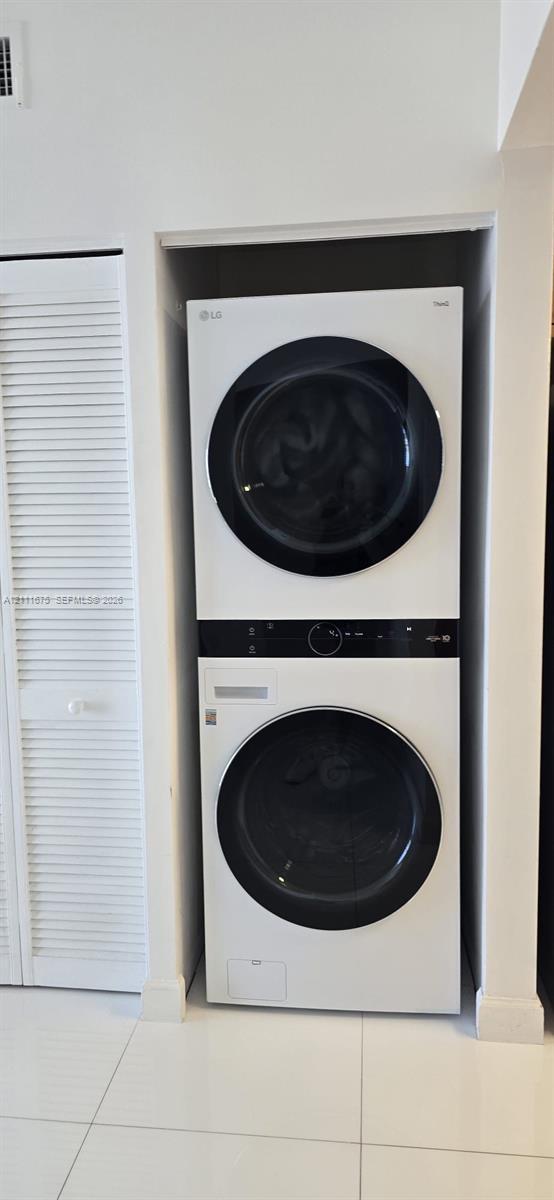 Stacked laundry unit featuring a white finish with front-loading glass-panel doors, complemented by white louvered closet doors and large-format tile 