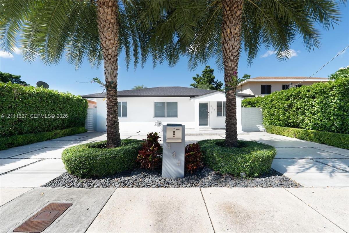 Single-story residence featuring a paved driveway with turf accents, mature palm trees, groomed privacy hedges, and a contemporary concrete mailbox pi