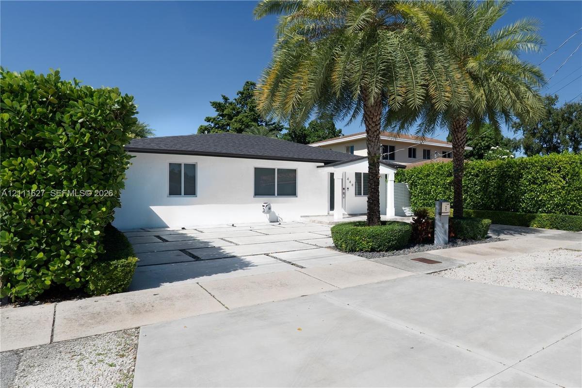 Single-story exterior featuring white stucco siding, a shingled roof, a geometric paver driveway, and mature palm trees