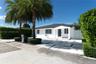 Single-story exterior featuring white stucco siding, a covered entryway with square columns, geometric concrete paver driveway, and mature palm trees