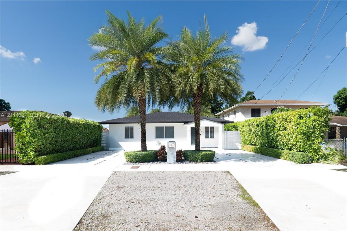 Single-story residence featuring a white exterior, hip roof, mature palm trees, and manicured privacy hedges