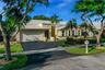 Single-story residence featuring a multi-gabled light-toned roof, an attached two-car garage, manicured landscaping with mature palm trees, and a pave