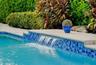 In-ground pool featuring a tiled water feature, blue glass mosaic waterline tile, light-toned concrete coping, and manicured perimeter landscaping