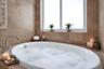 Oval soaking tub featuring a deck-mounted chrome faucet, set within stone-tiled wall surrounds and a window-integrated ledge