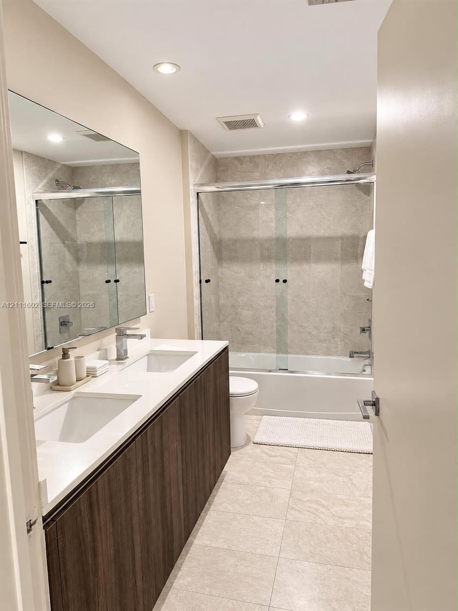 Dual-sink vanity with wood-finish cabinetry, quartz countertop, large wall-mounted mirror, and tub-shower combination featuring sliding glass doors an