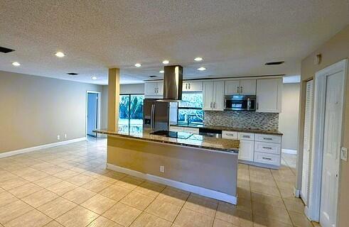 Kitchen featuring an island with a built-in cooktop and stainless steel vent hood, granite countertops, white cabinetry with silver hardware, stainles