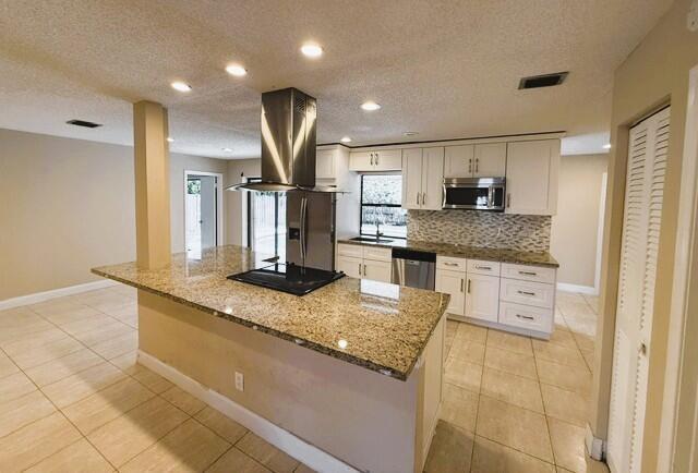 Kitchen featuring an island with a built-in cooktop and stainless steel vent hood, granite countertops, white cabinetry with silver hardware, stainles