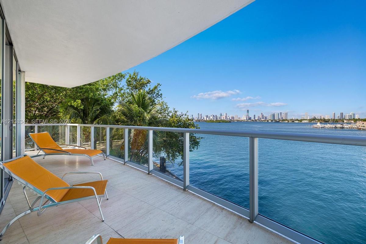 Covered balcony featuring stone flooring and a glass railing with metal framing, overlooking the waterfront and city skyline