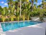 In-ground swimming pool with mosaic tile border, light-toned stone-finish patio, decorative masonry screen wall, and perimeter tropical landscaping
