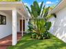 Courtyard entryway featuring a red brick floor, white stucco exterior walls, a natural wood-finish front door, and mature tropical landscaping