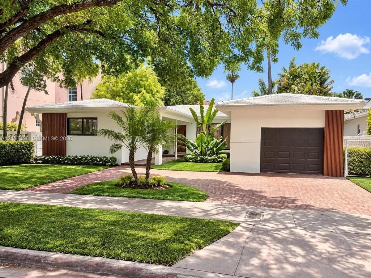 Single-story residence featuring a crisp white exterior with vertical wood-accented columns, a paved brick driveway, a dedicated garage, and mature la