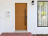 Wood-finish front door with a vertical matte black pull handle, white stucco exterior, exterior wall-mounted lighting, and herringbone-patterned brick