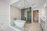 Enclosed wet room featuring a freestanding soaking tub, floor-to-ceiling glass paneling, dual-toned tiled walls, and a rainfall shower head