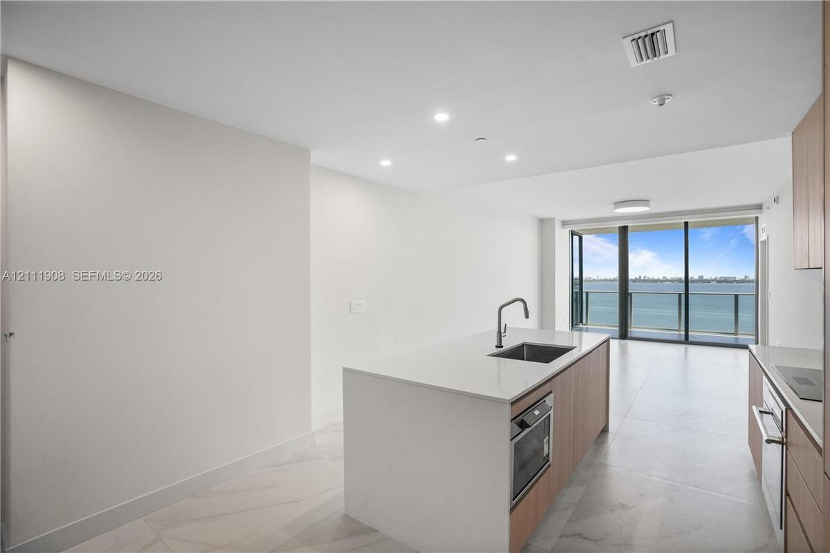 Kitchen island with stone-surface countertop, under-mount stainless steel sink, wood-finish cabinetry, floor-to-ceiling glass doors, and light-tone ti