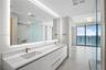 Dual-sink vanity with white cabinetry, quartz countertop, backlit mirror, and metallic fixtures featuring a glass-enclosed shower and floor-to-ceiling