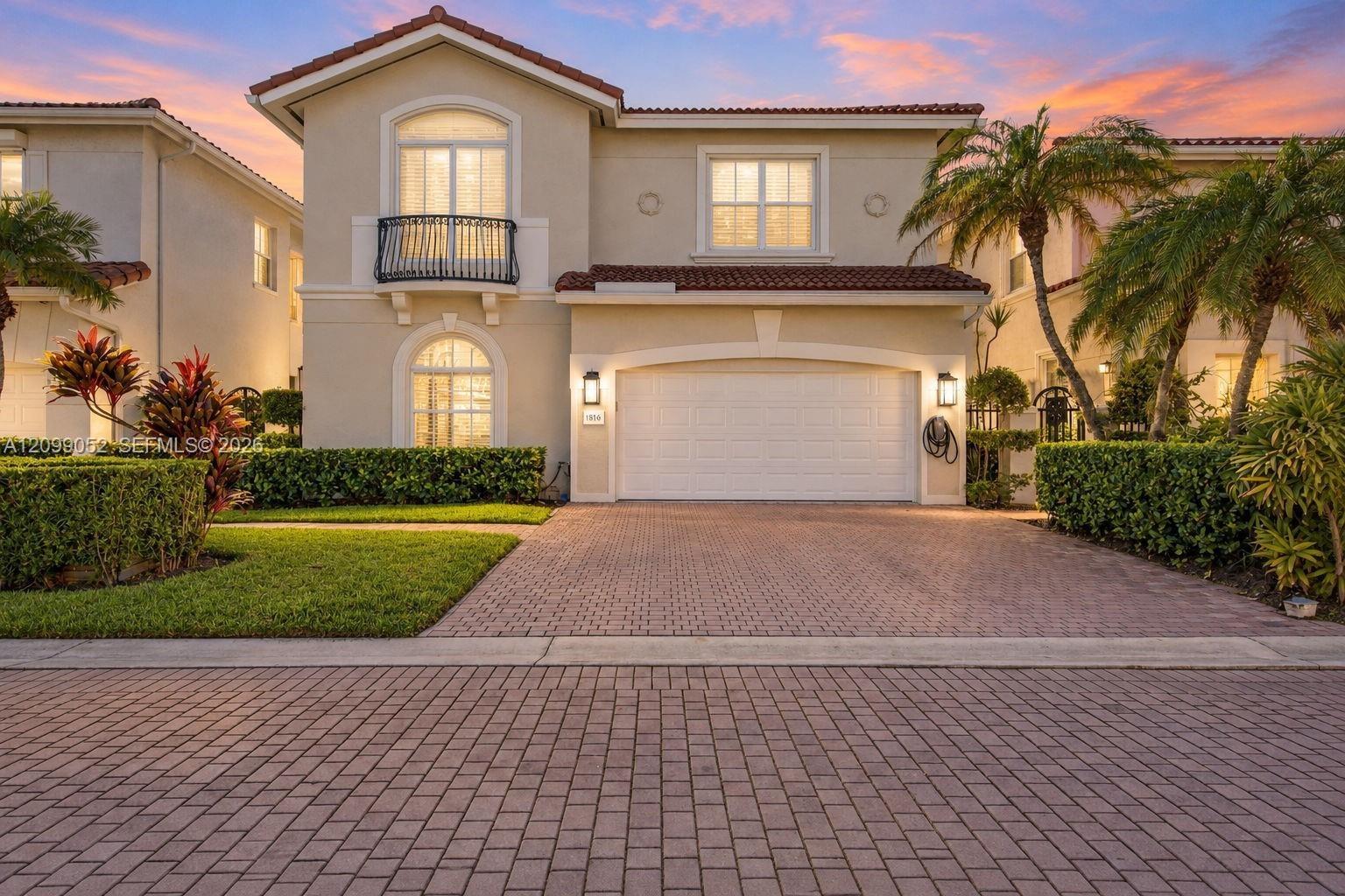 Stucco exterior featuring a terracotta tile roof, second-story balcony with metal railing, arched window detailing, and brick-paver driveway