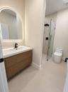 Floating vanity with wood-finish cabinetry, arched LED-backlit mirror, matte black faucet, and walk-in shower with glass panel