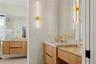 Floating wood-finish vanities with light stone countertops, integrated sinks, brushed gold faucets, and elongated vertical LED wall sconces