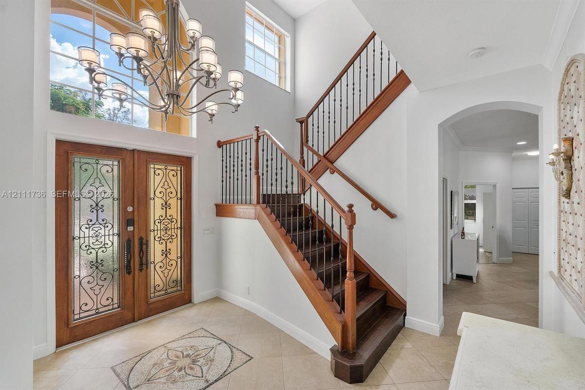Double-height foyer featuring a dual-door entryway with decorative ironwork inserts, a large multi-tiered chandelier, tile flooring with an inlaid med