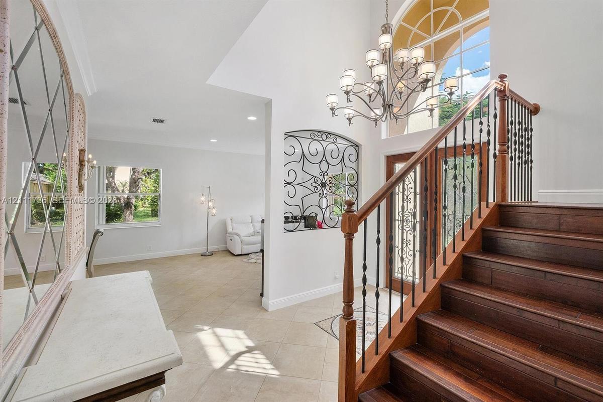Grand foyer featuring a high ceiling, wood-finish staircase with decorative metal balusters, arched window, wrought-iron wall accent, and multi-arm ch
