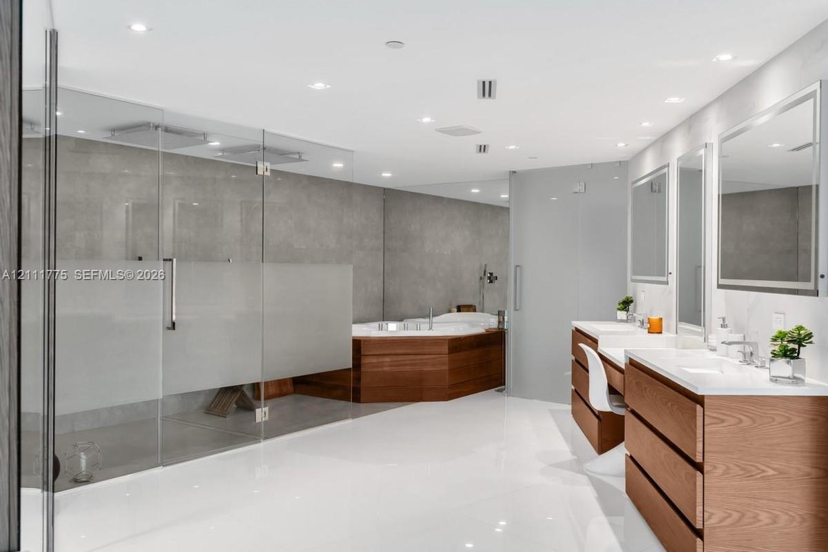Enclosed shower with glass paneling, wood-paneled soaking tub, dual wood-finish vanity cabinets with white countertops, and integrated mirror lighting